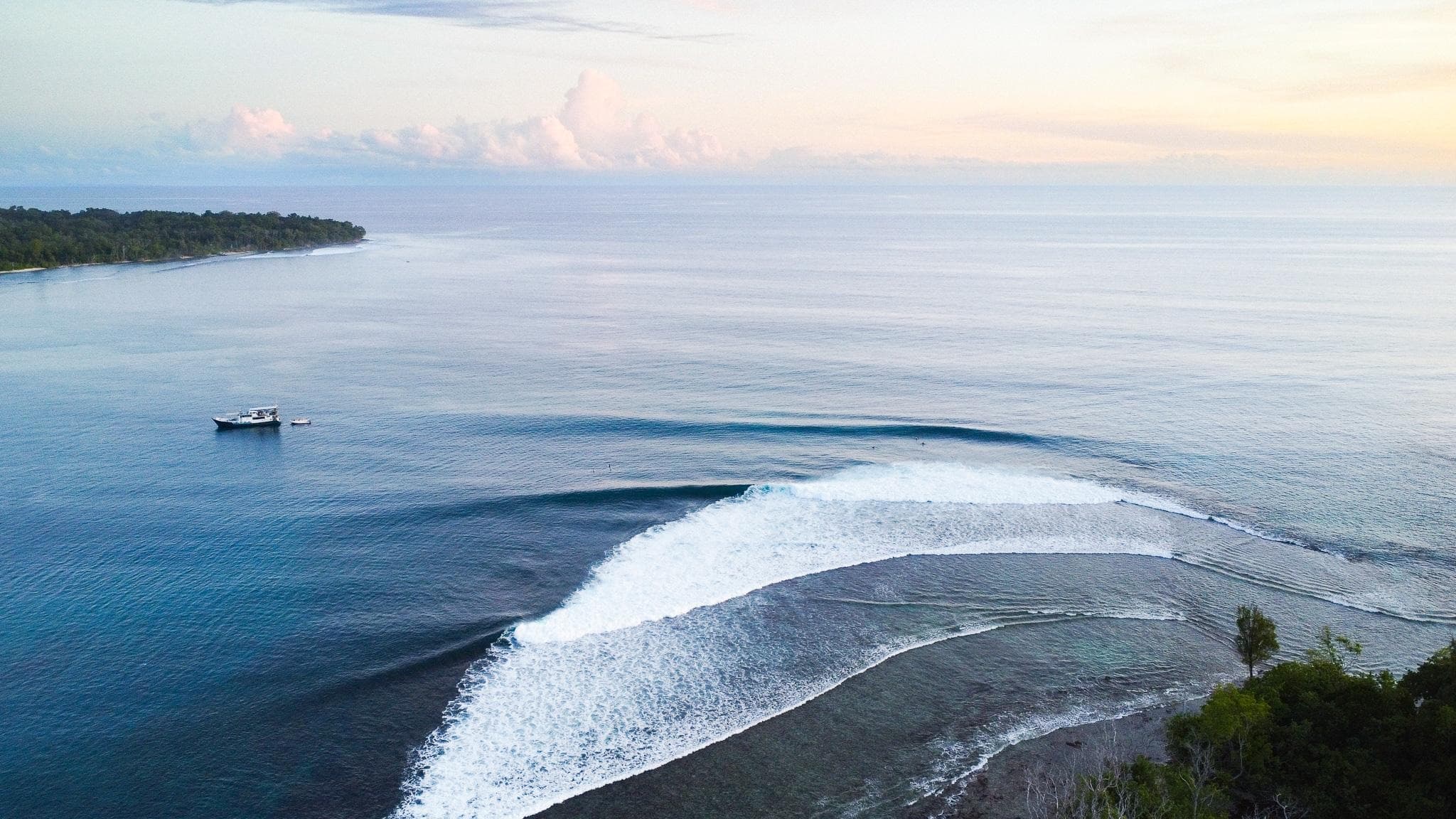 Drone shot of a reef-wrapped right-hander breaking at sunrise with a charter boat anchored offshore in the Mentawais.